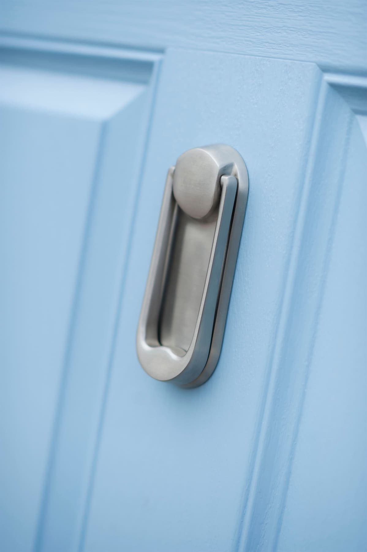 A satin chrome knocker face-fixed to the stile of a pale blue composite door, the grain of the moulding visible around it.